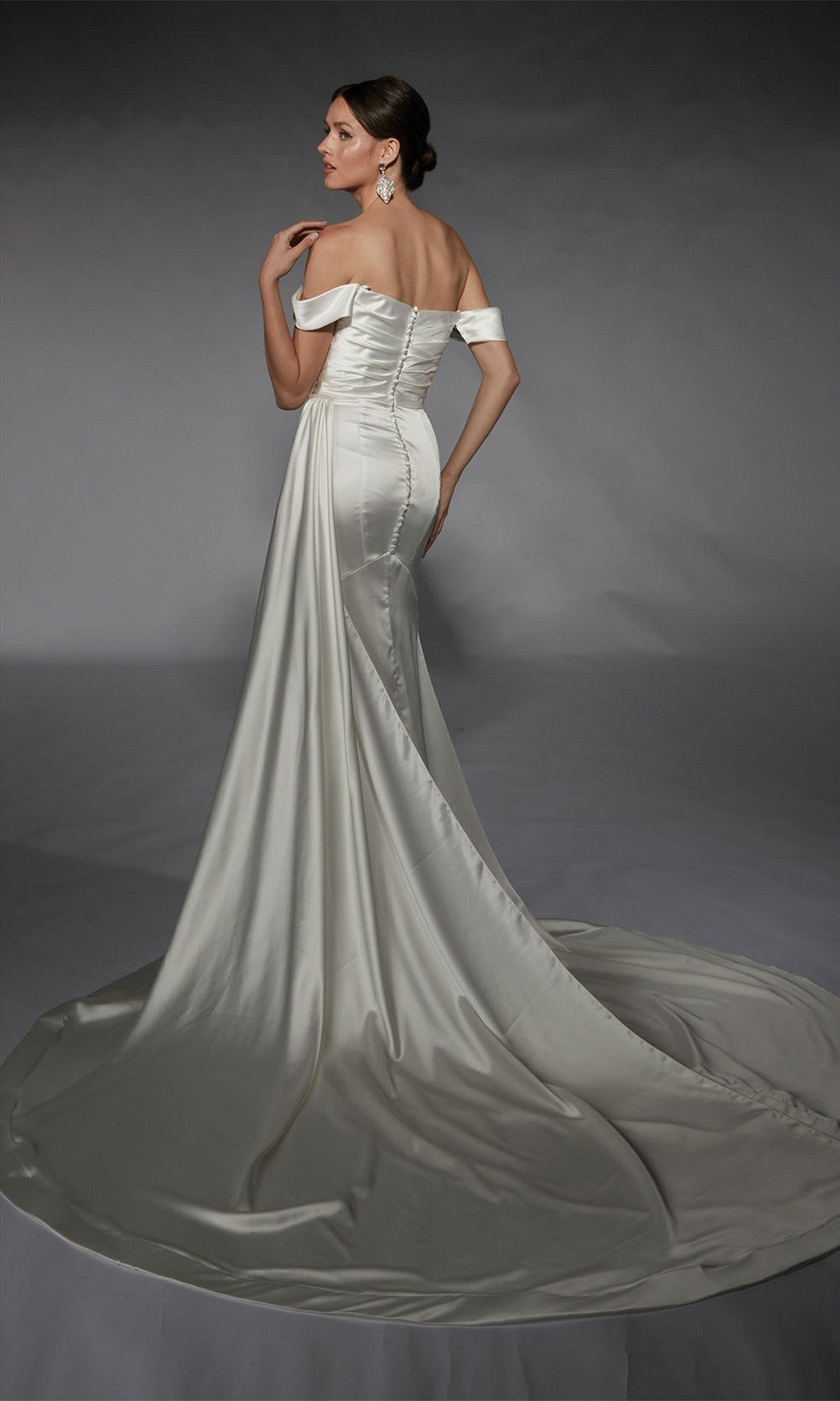 A woman stands facing away on a plain gray background, wearing the Alyce 7106 Long White Dress with an off-the-shoulder cowl neckline, white satin fabric, and a long train.